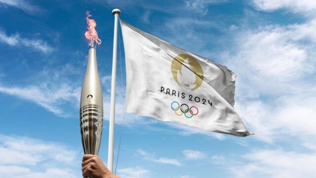 hand holding Olympic torch in front of “Paris 2024” flag